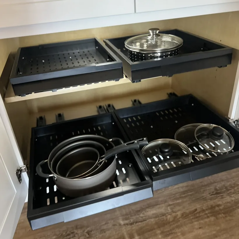 An open lower kitchen cabinet with two levels of pull-out sliding drawers, storing nesting dark gray pots with handles and glass lids on the lower shelf, and an empty tray alongside a steamer basket with glass lid on the upper shelf. White cabinet doors and wood-look flooring are visible.
