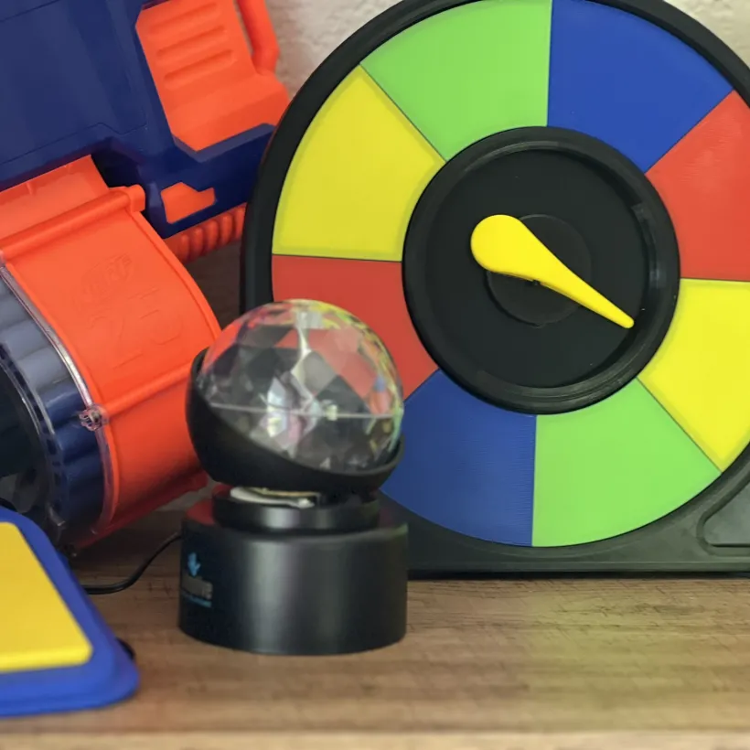 A colorful game room tabletop display featuring a Nerf-style foam dart blaster in blue and orange, a spinning color wheel with yellow, green, blue, and red sections and a yellow arrow pointer, a small rotating disco/light projector ball on a black base, and a yellow foam mat in the lower left corner.