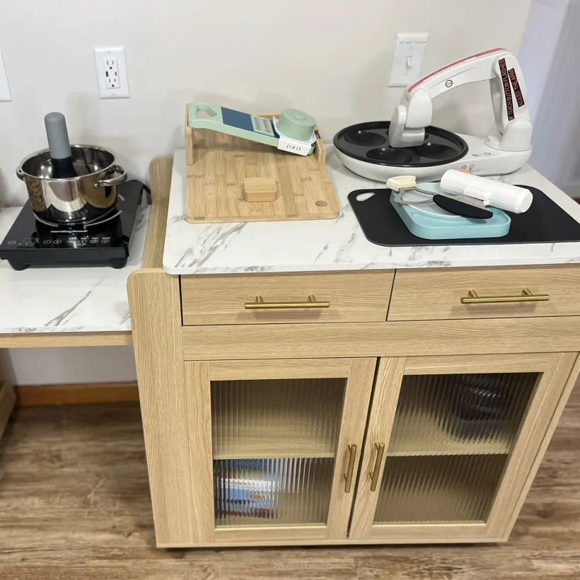 An adaptive kitchen training station with a wheelchair-accessible open-base table on the left holding a portable induction cooktop with a stainless steel pot and a wicker basket of kitchen tools. Beside it is a light wood cabinet with gold hardware and ribbed glass doors, topped with a bamboo cutting board with adaptive tools, a non-slip black cutting board with accessible utensils, and a white electric jar opener.