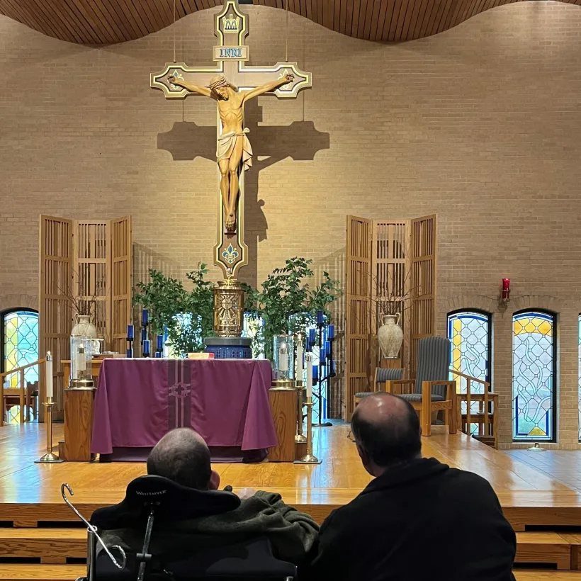 A Sunshine Communities resident and support staff member sit together in a church sanctuary during a faith service, facing the altar and crucifix in a peaceful moment of worship.