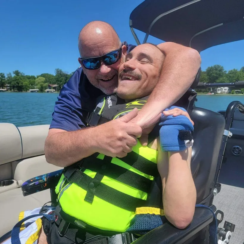 A father hugs his adult son while they enjoy a boat ride on a sunny lake, sharing a joyful moment together during a family outing supported by Sunshine Communities.