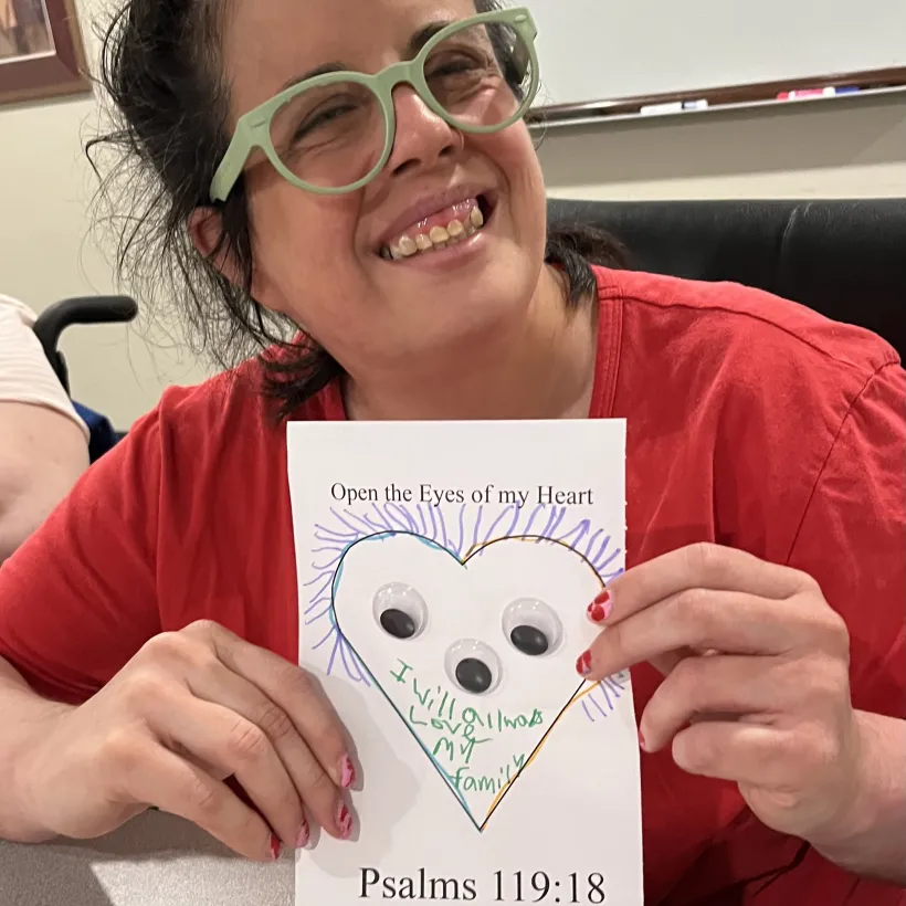 Participant at Sunshine Communities smiles while holding a completed Bible study coloring page featuring Psalm 1 19:18 during a Spiritual Life faith activity in Maumee, Ohio.