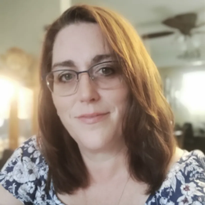Woman with shoulder-length brown hair and glasses wearing a blue and white floral top indoors.